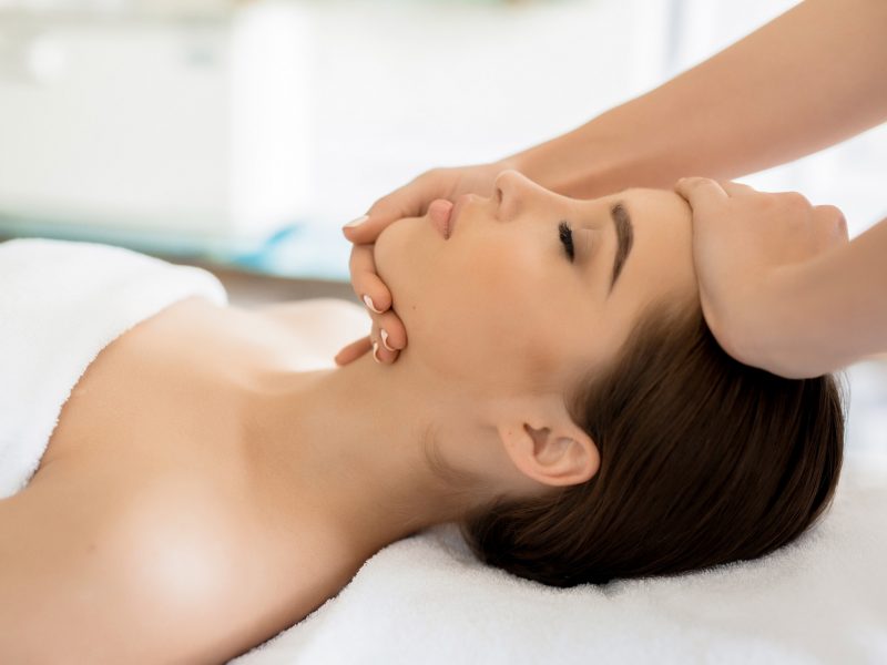 Hands of masseuse on forehead and chin of young client during procedure of spa facial massage