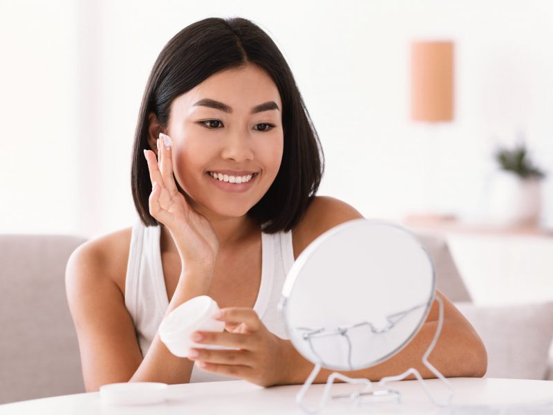 Female Skincare And Daily Routine Concept. Portrait Of Smiling Asian Lady Applying Moisturizer On Her Face Looking In The Mirror, Holding Hydration Cream For Healthy Facial Skin. Banner, Copy Space
